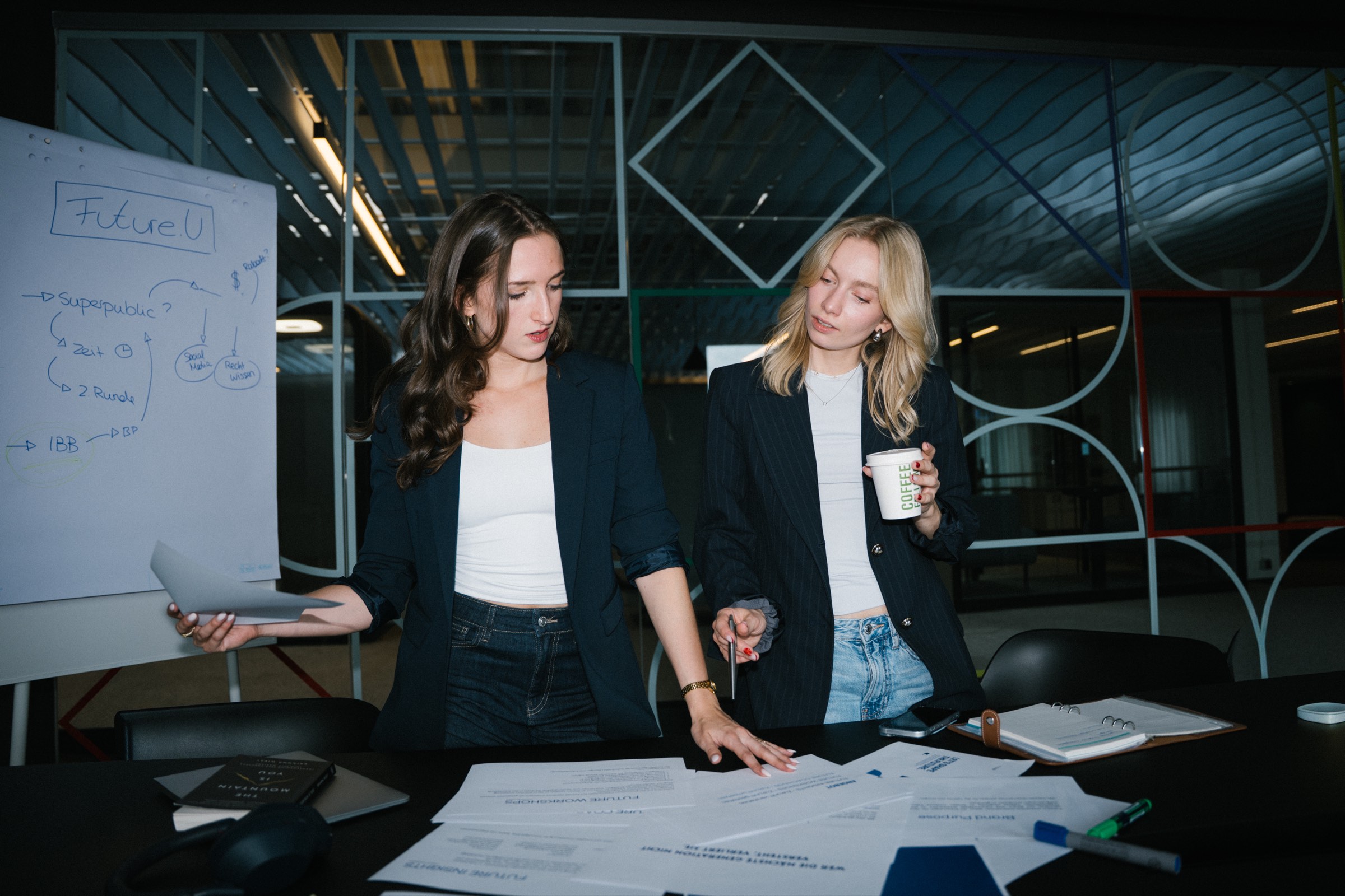 Johanna und Mavie von Future U arbeiten an einem Tisch voller Unterlagen, daneben ein Flipchart mit der Aufschrift Future.U